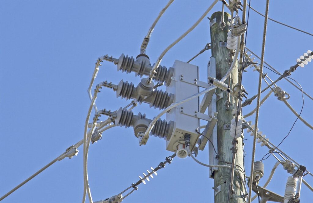 Electric Utility Infrastructure::A close-up view of electric utility equipment mounted on a wooden power pole against a clear blue sky.